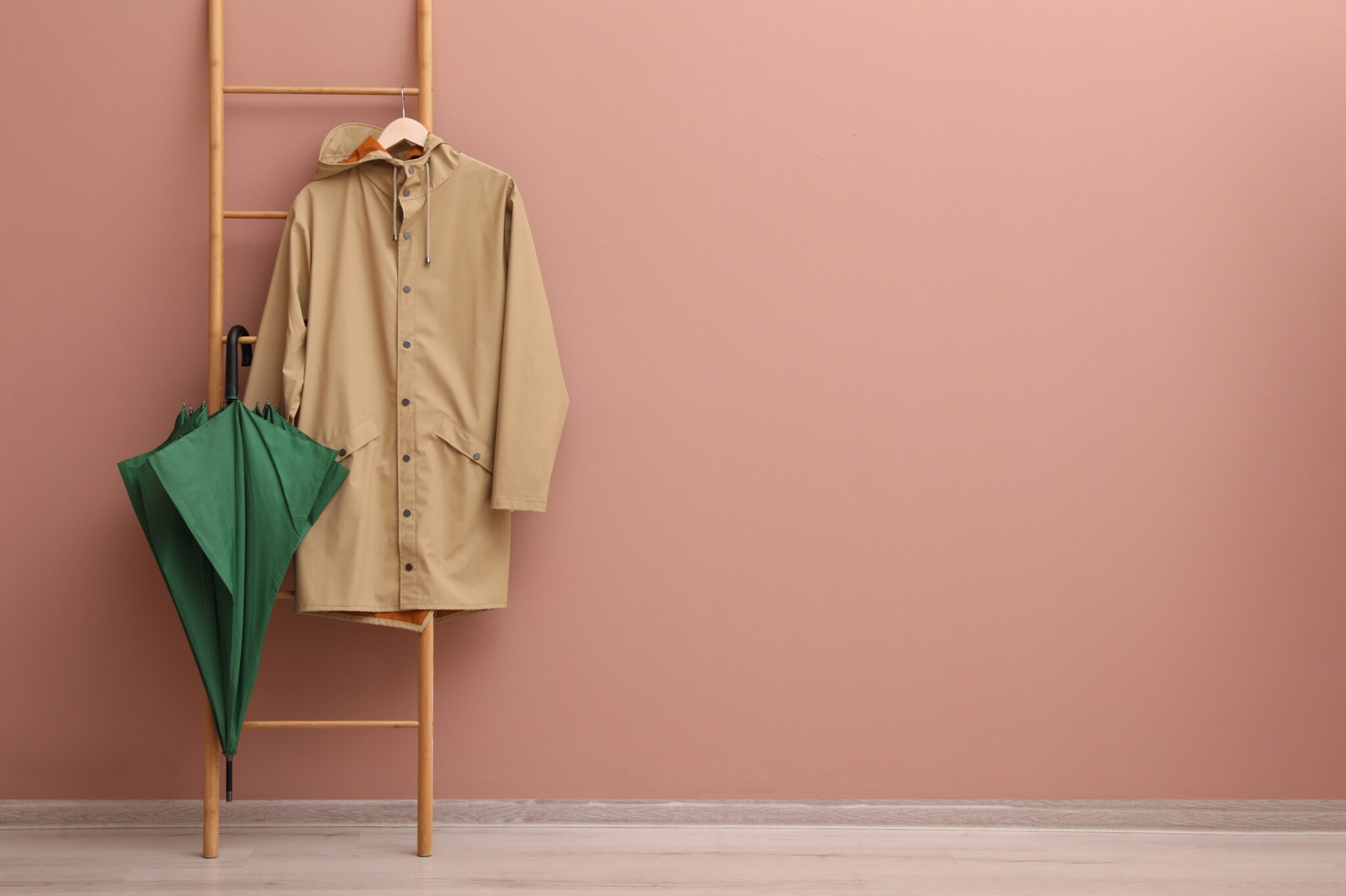Rainy day prep: Stylish green umbrella and raincoat on wooden ladder near beige wall indoors.