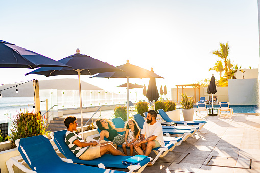 Family on vacation in a tropical location lounges by a luxurious pool, chatting and laughing