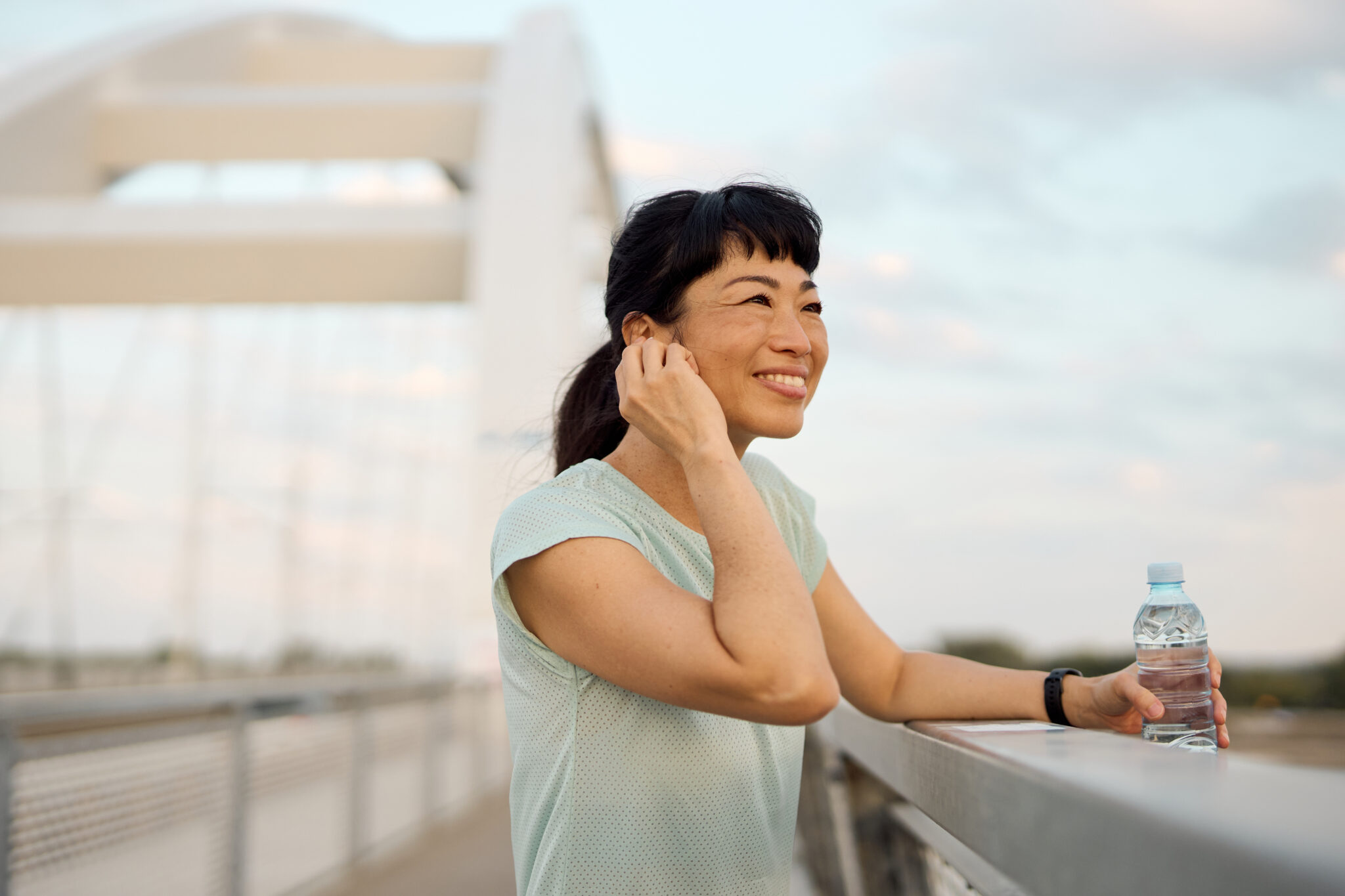 Woman wearing a hearing aid and workout clothes standing on a modern bridge, taking a break from exercising while holding a water bottle and looking cheerful
